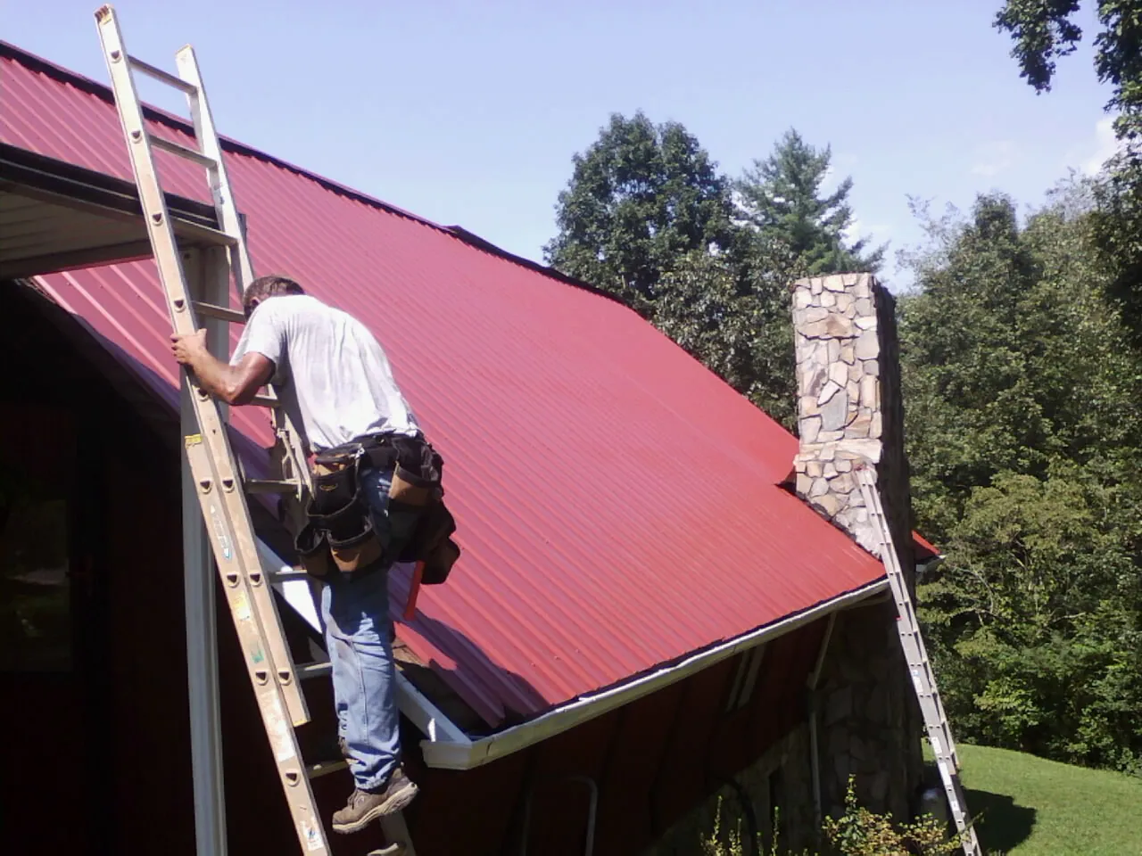 Roofer on a ladder beside a red metal roof and stone chimney — Soffit & Fascia Repair by Roofing Brothers in Starbuck